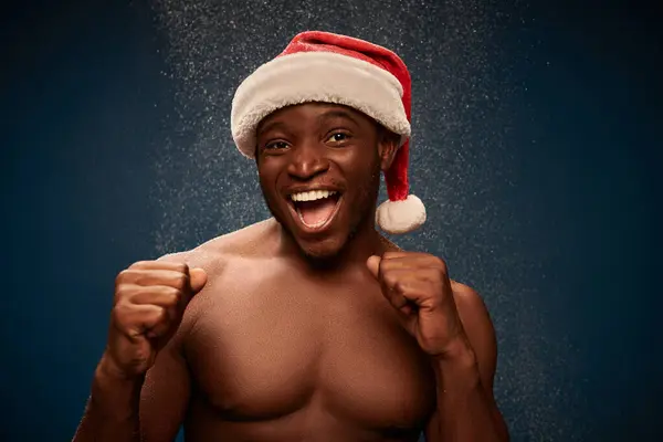 Eccitato uomo muscoloso afroamericano in cappello di Babbo Natale mostrando vincere gesto su sfondo blu navy innevato — Foto stock