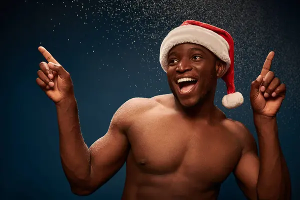 Aufgeregter athletischer afrikanisch-amerikanischer Mann mit Weihnachtsmütze, der mit dem Finger auf dunkel verschneite Kulisse zeigt — Stockfoto