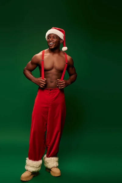 Cheerful muscular african american man in santa hat and red pants with suspenders on green backdrop — Stock Photo
