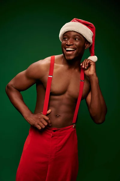 Fröhlicher, hemdloser afrikanisch-amerikanischer Mann mit Weihnachtsmütze und roter Hose, der vor grünem Hintergrund wegschaut — Stockfoto