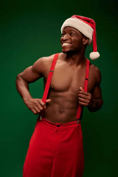 Excited shirtless african american guy in santa hat and red pants looking away on green background — Stock Photo
