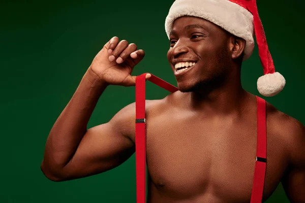 Athletic african american man in santa hat and suspenders smiling and looking away on green — Stock Photo