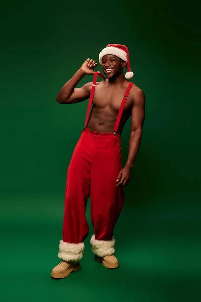 Muskulöser afrikanisch-amerikanischer Mann mit Weihnachtsmütze und roter Hose auf Hosenträgern lächelt in die Kamera auf grün — Stockfoto