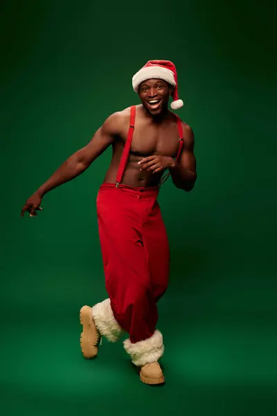 Excited athletic african american man in santa hat and red pants on suspenders posing on green — Stock Photo