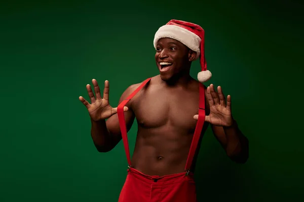 Smiley muscular african american guy in santa hat pants pulling suspenders and posing on green — Stock Photo