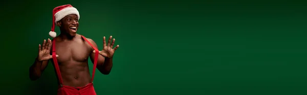 Веселый спортивный африканский американец, позирующий в шляпе Санты и штанах с подтяжками на зеленом, баннер — стоковое фото