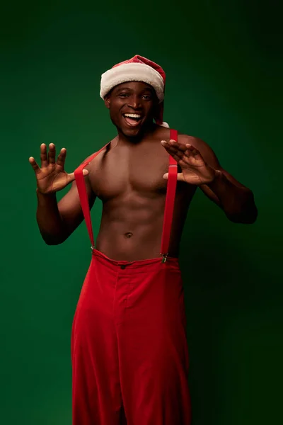 Shirtless muscular african american guy in santa hat pants pulling suspenders and smiling on green — Stock Photo