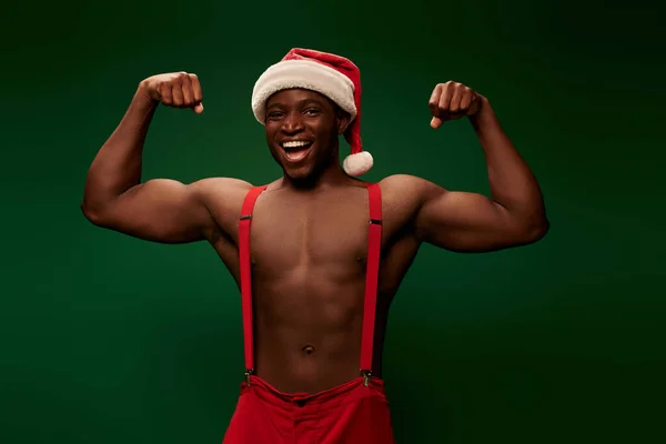 Fröhlicher und starker afrikanisch-amerikanischer Mann mit Weihnachtsmütze und Hose, der Muskeln vor grünem Hintergrund zeigt — Stockfoto