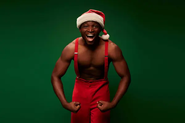 Energized and shirtless african american man in christmas hat and pants showing muscles on green — Stock Photo
