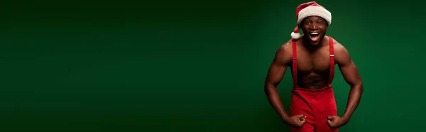 Homme afro-américain athlétique en chapeau de Noël et pantalon rouge montrant les muscles sur vert, bannière — Photo de stock