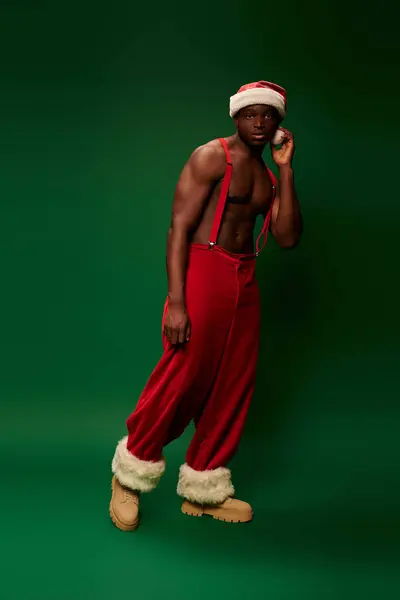Thoughtful and sportive african american man in santa hat and red pants looking at camera on green — Stock Photo