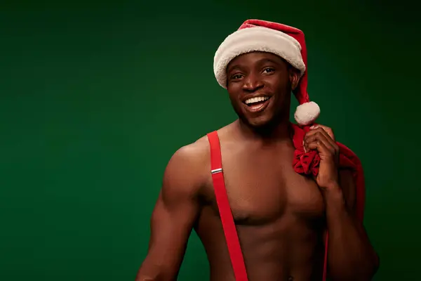 Muscular african american man in santa hat holding christmas bags and looking at camera on green — Stock Photo