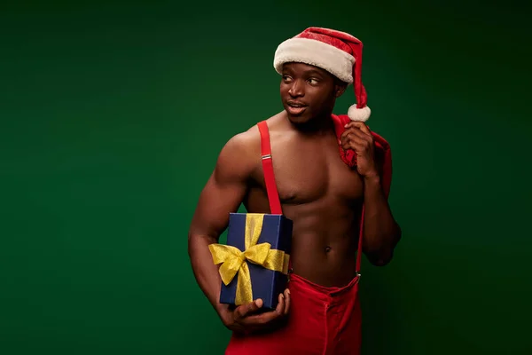 Homem americano africano sem camisa em chapéu de santa e calças vermelhas com caixa de presente e saco de natal em verde — Fotografia de Stock
