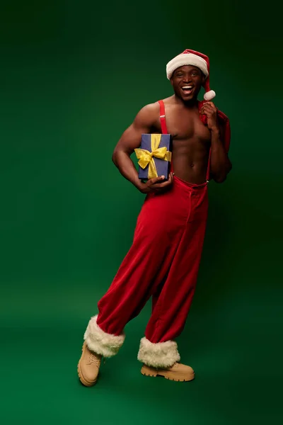 Overjoyed shirtless african american man in santa hat holding christmas bag and gift box on green — Stock Photo