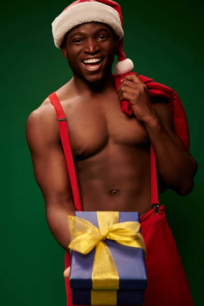 Joyful shirtless african american man in santa hat with christmas bag showing gift box on green — Stock Photo
