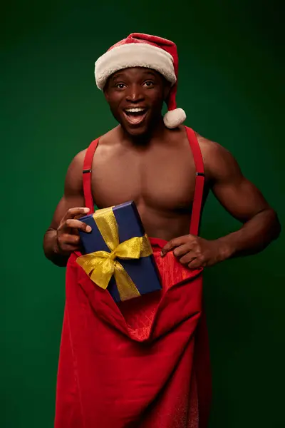 Excited athletic african american man in santa hat with christmas bag and gift box on green backdrop — Stock Photo