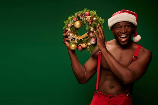 Joyous athletic african american man in santa hat holding christmas wreath with baubles on green — Stock Photo