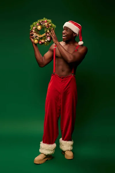 Amazed african american man in santa hat and red pants looking at shiny christmas wreath on green — Stock Photo