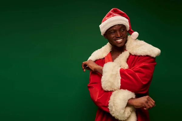Веселый африканский американец в праздничном костюме Санта Клауса, смотрящий в сторону на зеленом фоне — стоковое фото