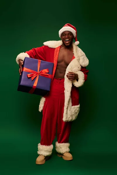 Задоволений афроамериканський чоловік у костюмі Санти Клауса, що стоїть з величезною подарунковою коробкою на зеленому тлі — стокове фото