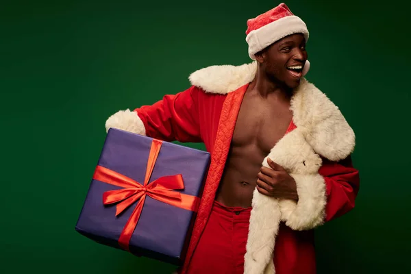 Щасливий афроамериканський чоловік у різдвяному костюмі на без сорочки, що тримає величезний подарунок на зеленому — стокове фото