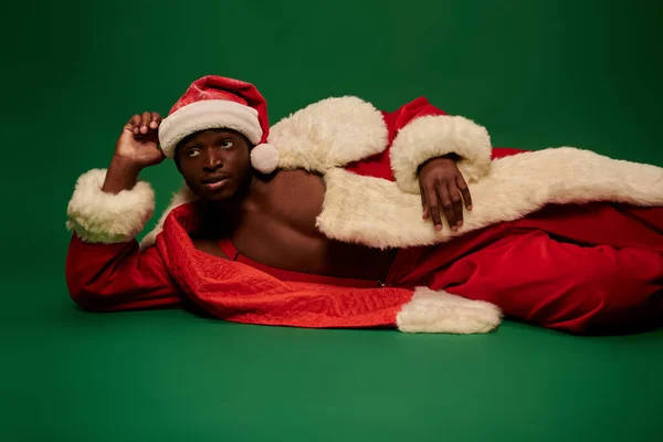 Sexy afrikanisch-amerikanischer Mann im Weihnachtsmann-Kostüm auf hemdslosem Körper liegend und wegschauend auf grün — Stockfoto