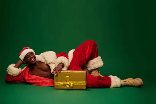Quente afro americano cara no santa traje deitado perto brilhante dom caixa e sorrindo no verde — Fotografia de Stock