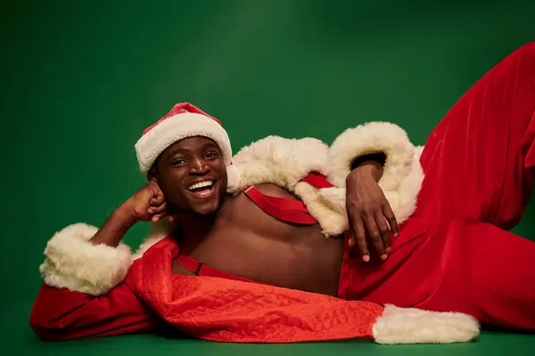 African american man with radiant smile lying down in santa costume on shirtless body on green — Stock Photo