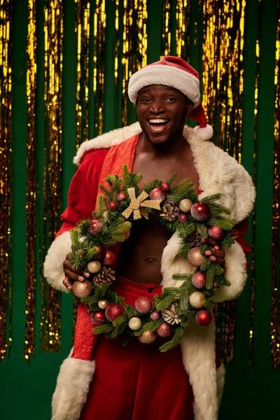 Heureux homme afro-américain en costume de Père Noël avec couronne de Noël près de tinsel d'or sur vert — Photo de stock