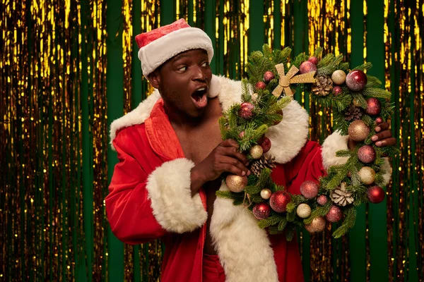 Entusiasta uomo afro-americano in costume da Babbo Natale guardando ghirlanda di Natale su sfondo dorato fili di lame — Foto stock