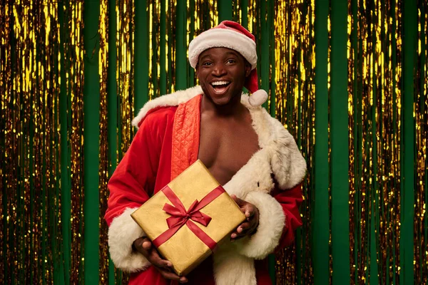 Heureux homme afro-américain en costume de Père Noël sur corps torse nu avec boîte cadeau brillant près tinsel doré — Photo de stock