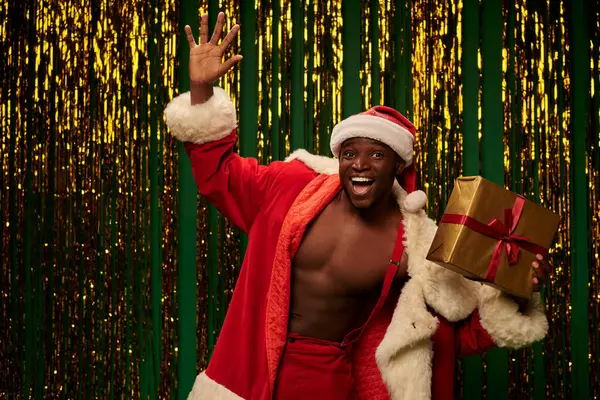 Jolly african american man in christmas costume holding present and waving hand on shiny backdrop — Stock Photo