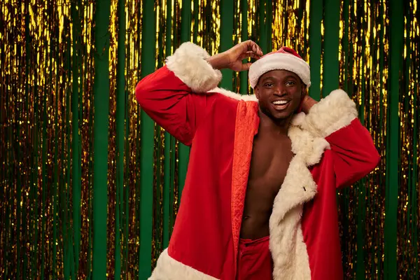 Gai afro-américain gars posant en costume de Père Noël sur corps torse nu près tinsel brillant sur vert — Photo de stock