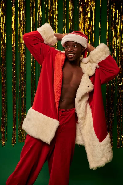 Sexy and shirtless african american guy in santa costume with hands behind head near golden tinsel — Stock Photo