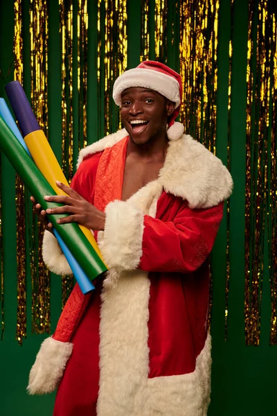Excited african american man in santa costume with colorful wallpapers on green backdrop with tinsel — Stock Photo