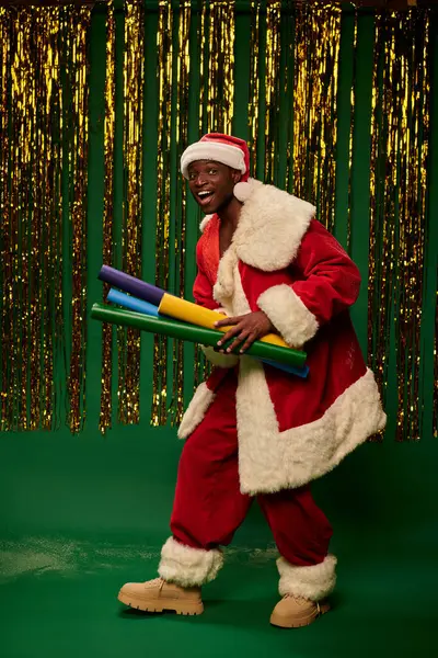 Joyful african american guy in santa costume with colorful paper rolls on green backdrop with tinsel — Stock Photo