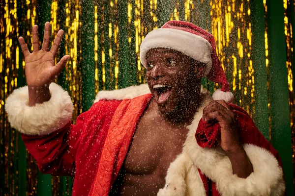 Homem americano africano em traje de santa com saco de natal acenando mão em pano de fundo nevado e brilhante — Fotografia de Stock