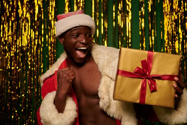 Super alegre homem afro-americano em traje de santa com presente de Natal sob neve em estúdio brilhante — Fotografia de Stock