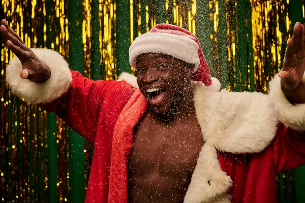 Excited african american man in santa costume on shirtless body gesturing on shiny snowy backdrop — Stock Photo