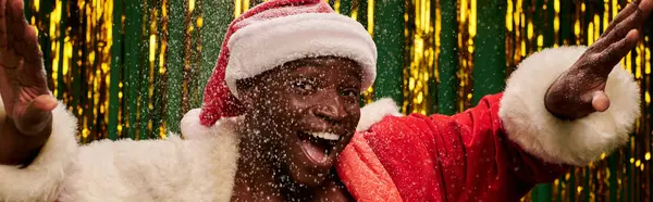 Cheerful african american man in santa costume gesturing under snowfall on golden backdrop, banner — Stock Photo