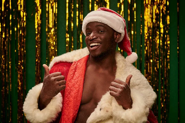 Hot dark skinned guy in santa costume on shirtless body showing thumbs up on christmas backdrop — Stock Photo