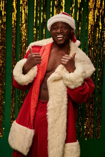 Sexy afro-américain guy en costume de Père Noël sur corps torse nu montrant pouces sur fond brillant — Photo de stock