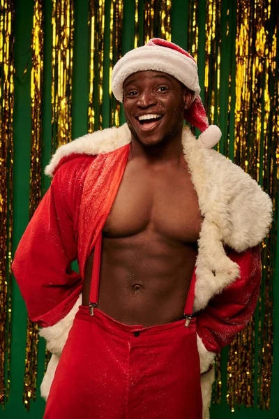 Sexy sportive african american man in santa costume on shirtless body near golden tinsel on green — стоковое фото