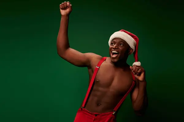 Збуджений без сорочки афроамериканський чоловік у різдвяному капелюсі та штанах радіє та кричить на зеленому — стокове фото