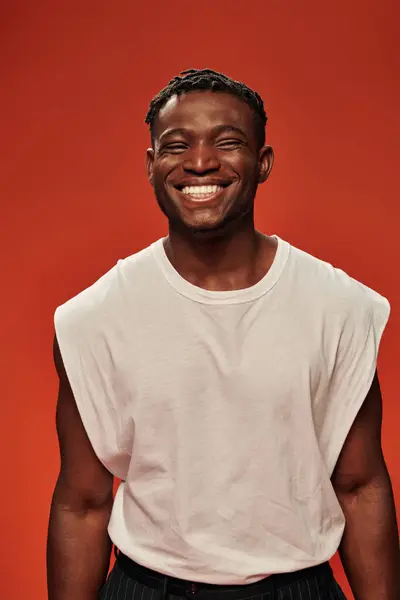 Homme afro-américain heureux et sportif en débardeur blanc souriant à la caméra sur rouge, modèle masculin moderne — Photo de stock