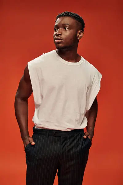 Stylish african american man in white tank top and black pants standing with hands in pockets on red — Stock Photo