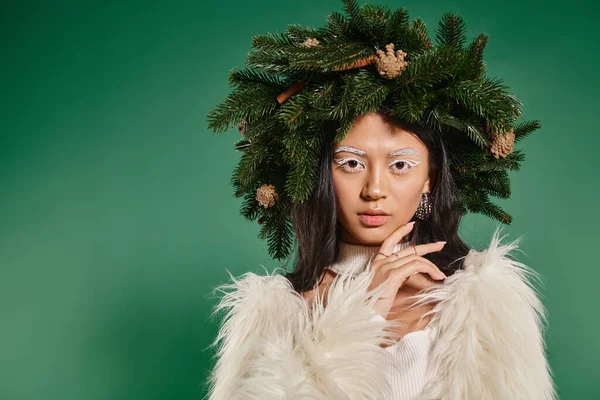 Attractive woman in faux fur jacket and natural wreath on head looking at camera on green backdrop — Stock Photo