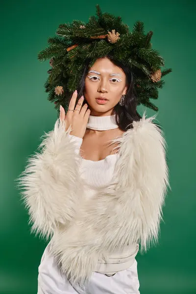 Asian woman in winter faux fur jacket and natural wreath on head looking at camera on green backdrop — Stock Photo