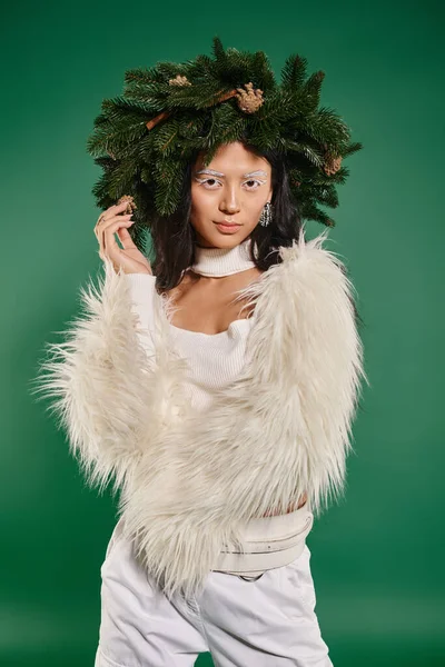 Winterkonzept, asiatische Frau mit weißem Make-up und trendigem Outfit posiert im Kranz vor grünem Hintergrund — Stockfoto