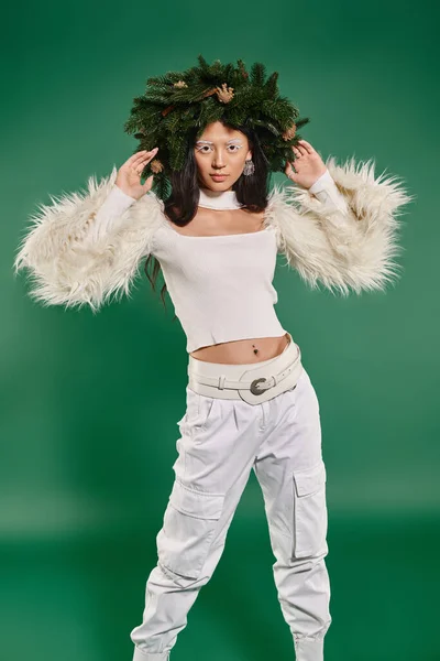 Winter concept, pretty woman with white makeup and trendy outfit posing in wreath on green backdrop — Stock Photo